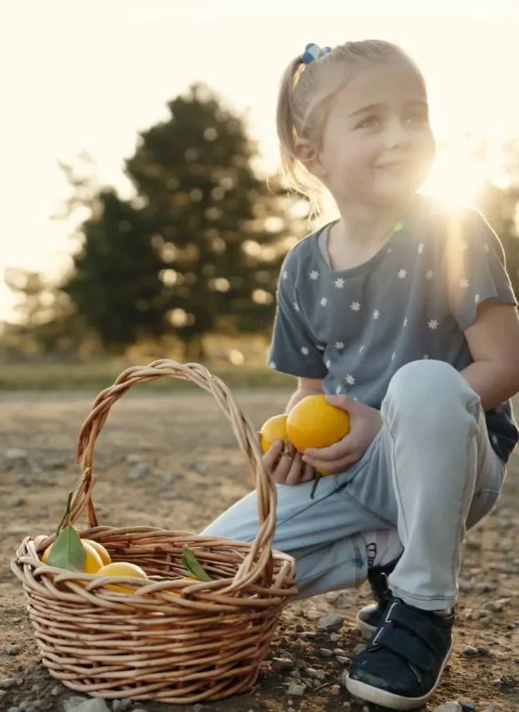 A cheerful girl sits on the ground, holding a basket brimming with vibrant lemons, enjoying her time outdoors.