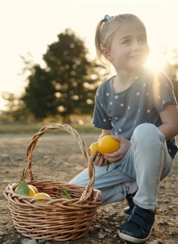 A cheerful girl sits on the ground, holding a basket brimming with vibrant lemons, enjoying her time outdoors.